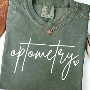 Puede incluir: Camiseta verde oliva con la palabra "optometry" en escritura blanca y un pequeño corazón. Un collar dorado con un colgante descansa sobre la camiseta. La camiseta tiene una etiqueta Comfort Colors.