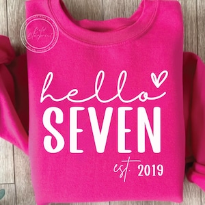 May include: A bright pink sweatshirt with the words "hello SEVEN" in white script and block letters, with a small heart above the "N". Below the word "SEVEN" is the text "est. 2019". The sweatshirt is a crew neck style.