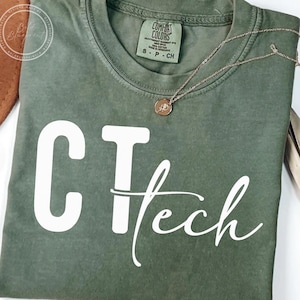 Peut inclure: T-shirt vert olive avec le texte "CT Tech" en blanc. Un collier doré avec un petit pendentif repose sur le t-shirt. Le t-shirt porte une étiquette Comfort Colors.
