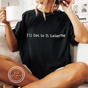 May include: Black t-shirt with the text "I'll Get to It Later™" in white. A white coffee mug is held in the left hand. The shirt is part of a clothing collection.