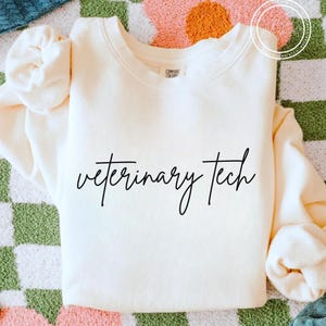 May include: Cream-colored sweatshirt with the words "veterinary tech" in black script. The sweatshirt has a crew neck and long sleeves. The background is a mix of colors and patterns.