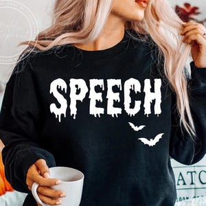 May include: Black sweatshirt with the word "SPEECH" in dripping white letters and two white bat silhouettes. The sweatshirt is a Halloween-themed design.