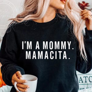 May include: Black sweatshirt with the text "I'M A MOMMY. MAMACITA." in white block letters. The sweatshirt is a crew neck style. A person is holding a white mug. The background is blurred.
