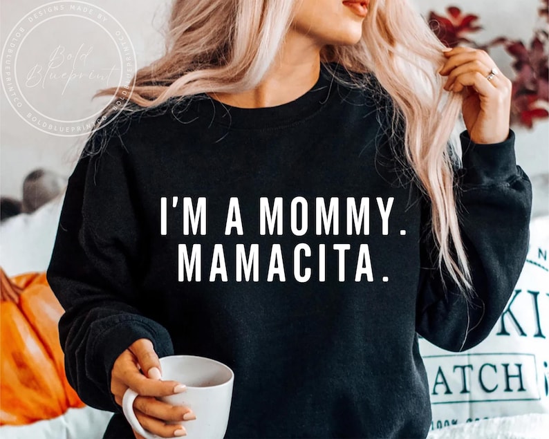 May include: Black sweatshirt with the text "I'M A MOMMY. MAMACITA." in white block letters. The sweatshirt is a crew neck style. A person is holding a white mug. The background is blurred.