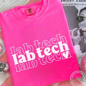 Op de afbeelding: Een felroze t-shirt met de tekst "lab tech" in het wit, verticaal gestapeld. Een klein wit hartje is toegevoegd. Het shirt is opgevouwen en gepresenteerd op een roze en wit geruit oppervlak.