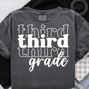 May include: A dark gray long-sleeve t-shirt with the words "third grade" in large, white, stylized lettering. The shirt has a crew neck and a relaxed fit. The text is stacked vertically, with "third" repeated twice.