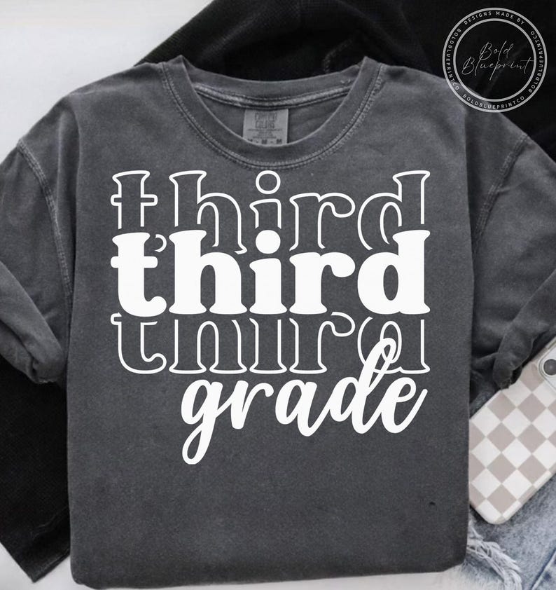 May include: A dark gray long-sleeve t-shirt with the words "third grade" in large, white, stylized lettering. The shirt has a crew neck and a relaxed fit. The text is stacked vertically, with "third" repeated twice.
