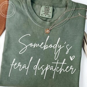 以下が含まれることがあります： オリーブグリーンのTシャツ。白の筆記体で「Somebody's feral dispatcher」と書かれています。金色のネックレスがTシャツの上にかけられています。Comfort ColorsブランドのSサイズTシャツです。