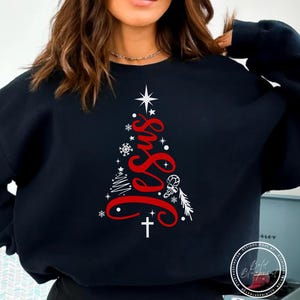 May include: Black sweatshirt featuring a red Christmas tree design with the word "Jesus" in a stylized font. The tree includes snowflakes, a star, and a cross. The design is printed on the front of the sweatshirt.