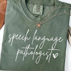 May include: An olive green t-shirt with the words "speech language pathologist" in white script. A gold-colored necklace with a small pendant rests on the shirt. The shirt has a crew neck and short sleeves.