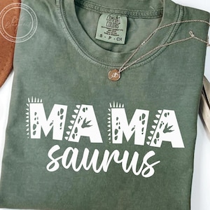 May include: Green t-shirt with white text that reads "Mama saurus" with a dinosaur-inspired design around the letters.