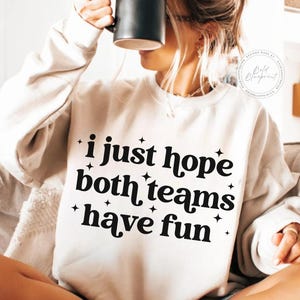 May include: Cream-colored sweatshirt with the text "i just hope both teams have fun" in black. The text is surrounded by small star graphics. The sweatshirt is a casual, comfortable style.