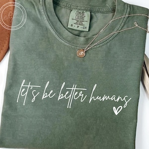 May include: An olive green t-shirt with the phrase "let's be better humans" in white script, accompanied by a small heart. A gold necklace with a pendant rests on the shirt. The shirt is folded neatly.
