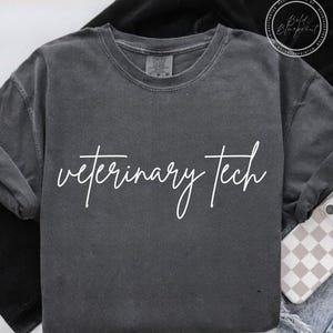 May include: Dark gray long-sleeve shirt with the words "veterinary tech" in white script. The shirt is made of a soft material and has a crew neck. A phone with a checkered case is visible.