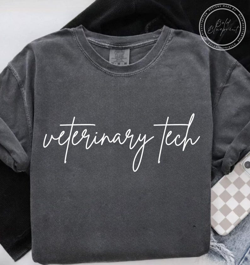 May include: Dark gray long-sleeve shirt with the words "veterinary tech" in white script. The shirt is made of a soft material and has a crew neck. A phone with a checkered case is visible.