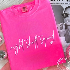 Könnte beinhalten: Ein leuchtend pinkfarbenes T-Shirt mit Rundhalsausschnitt und dem weißen Schriftzug "night shift squad". Ein kleines Herz folgt dem Wort "squad". Das Shirt ist einfarbig und aus weichem Material.