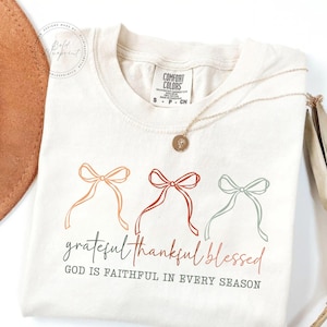 May include: Cream-colored t-shirt with the words "grateful thankful blessed" and the phrase "GOD IS FAITHFUL IN EVERY SEASON" printed in a script font. The shirt features three bow designs in orange, red, and blue-green. A gold necklace is draped across the shirt.