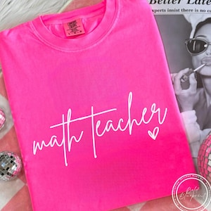 May include: Bright pink t-shirt with the words "math teacher" in white script. A small white heart is next to the text. The shirt is folded and appears to be made of a soft material.