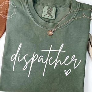 Peut inclure: T-shirt vert olive avec le mot "dispatcher" en écriture blanche et un petit cœur. Un collier doré avec un pendentif repose sur le t-shirt. Le t-shirt a une étiquette Comfort Colors.