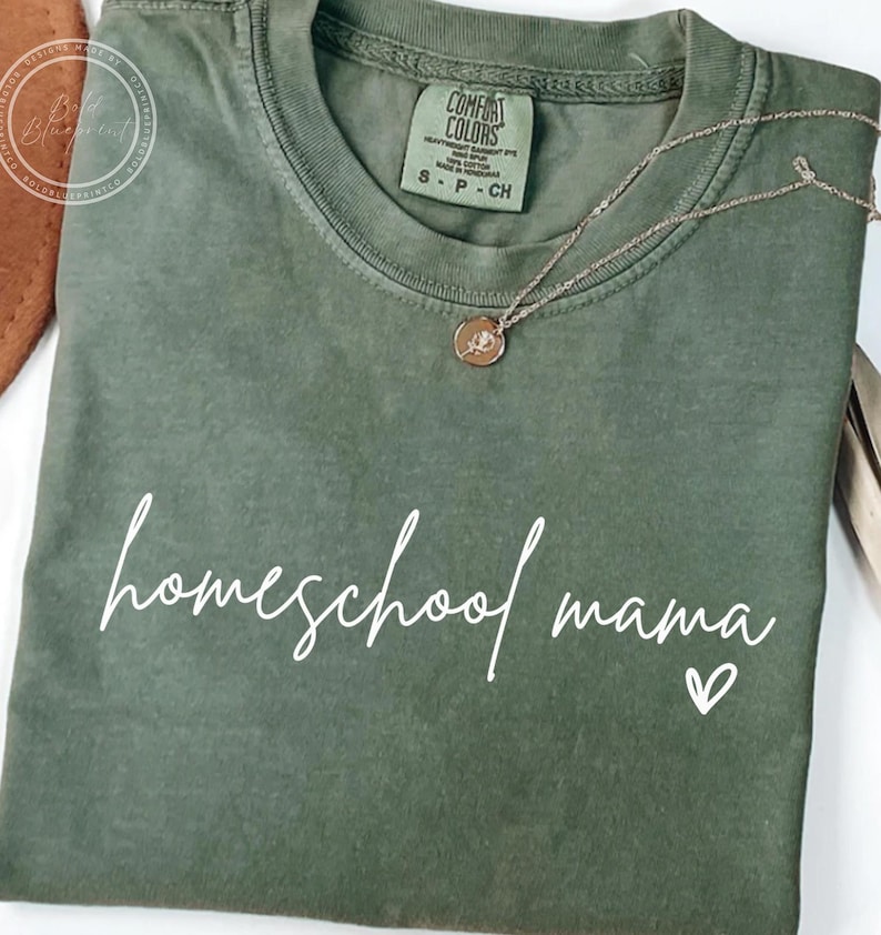May include: Olive green t-shirt with the words "homeschool mama" in white script and a small heart. A gold necklace with a pendant rests on the shirt. The shirt is folded and has a Comfort Colors tag.