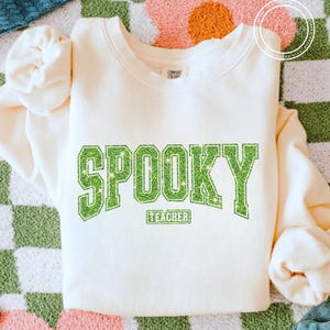 May include: Cream-colored sweatshirt with the word "SPOOKY" in large, glittery green letters. Below the word "SPOOKY" is the word "TEACHER" in a smaller, rectangular green box. The sweatshirt has a crew neck and long sleeves.