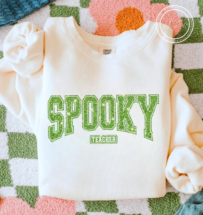 May include: Cream-colored sweatshirt with the word "SPOOKY" in large, glittery green letters. Below the word "SPOOKY" is the word "TEACHER" in a smaller, rectangular green box. The sweatshirt has a crew neck and long sleeves.