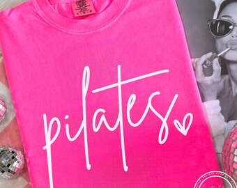 May include: A bright pink t-shirt with the word "pilates" in white script, with a small heart at the end. The shirt is folded and lies on a pink and white checkered surface. The Comfort Colors brand tag is visible.