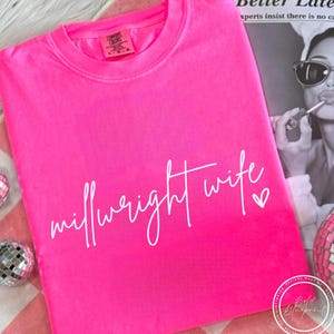 Peut inclure: Un t-shirt rose vif plié, avec l'inscription blanche "millwright wife" et un cœur. Le col du t-shirt porte une étiquette. Des boules disco et un magazine sont en arrière-plan.