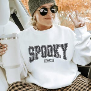 May include: White sweatshirt with the word "SPOOKY" in a glittery, block letter design. Below the word "SPOOKY" is the word "TEACHER". The sweatshirt is worn with a gray beanie, sunglasses, and a gold chain necklace.