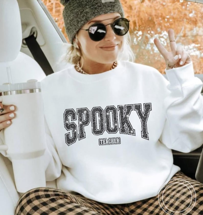 May include: White sweatshirt with the word "SPOOKY" in a glittery, block letter design. Below the word "SPOOKY" is the word "TEACHER". The sweatshirt is worn with a gray beanie, sunglasses, and a gold chain necklace.