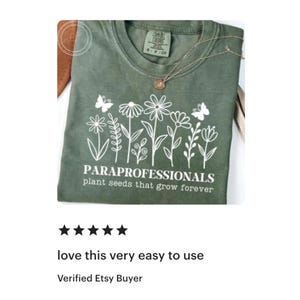 May include: Olive green t-shirt with white floral design and text "PARAPROFESSIONALS plant seeds that grow forever." A gold necklace with a pendant is draped on the shirt. The shirt is folded on a brown surface.