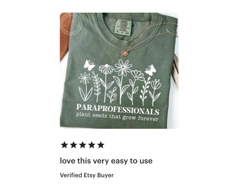 May include: Olive green t-shirt with white floral design and text "PARAPROFESSIONALS plant seeds that grow forever." A gold necklace with a pendant is draped on the shirt. The shirt is folded on a brown surface.