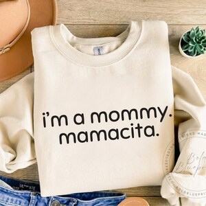 May include: Cream-colored sweatshirt with the text "i'm a mommy. mamacita." in black. The sweatshirt is folded, with a brown hat and blue jeans visible in the background. A small succulent plant in a white pot is also present.