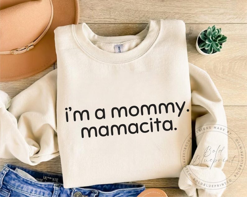 May include: Cream-colored sweatshirt with the text "i'm a mommy. mamacita." in black. The sweatshirt is folded, with a brown hat and blue jeans visible in the background. A small succulent plant in a white pot is also present.