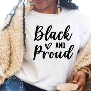Peut inclure: Un t-shirt blanc avec le texte "Black and Proud" en lettres cursives noires avec un symbole de cœur entre les mots.