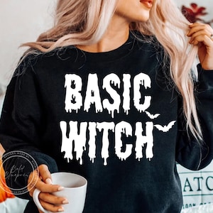 May include: A black long-sleeve crewneck sweatshirt featuring the white text "BASIC WITCH" in a dripping, horror-inspired font. Two small white bat silhouettes are positioned to the right of the word "BASIC" on the front of the garment.