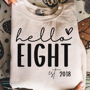 Könnte beinhalten: Beiges Sweatshirt mit den schwarzen Worten "hello EIGHT" in Schreib- und Blockschrift, mit einem kleinen Herz über dem Wort "EIGHT". Darunter steht "est. 2018".