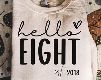 May include: A beige sweatshirt with the words "hello EIGHT" in black script and block letters, with a small heart above the word "EIGHT". Below the word "EIGHT" is the text "est. 2018".