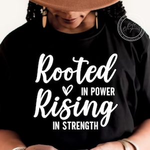 Pode incluir: Camiseta preta com o texto branco "Rooted in Power Rising in Strength". A camisa apresenta um símbolo de coração entre "Rooted" e "in Power". A camisa é usada por uma pessoa que usa um chapéu marrom e brincos de ouro.