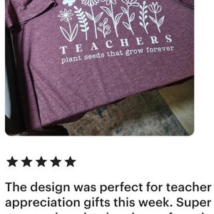 May include: Maroon t-shirt with a white floral design and the text "TEACHERS plant seeds that grow forever." The shirt has short sleeves and a crew neck. The design includes flowers and butterflies.