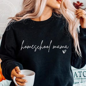 May include: Black sweatshirt with the words "homeschool mama" in white script, with a small heart. The sweatshirt is a crew neck style. A person is holding a white mug.