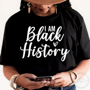 May include: A black t-shirt with white text that reads "I am Black History" with a small heart symbol. The shirt is being worn by a person with brown skin and a brown hat.