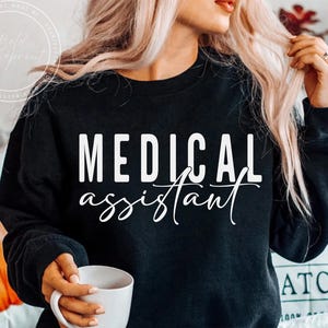 Può includere: Felpa nera con le parole "MEDICAL assistant" in bianco in carattere a blocchi e corsivo. La felpa è a girocollo. Una tazza bianca è tenuta in mano.