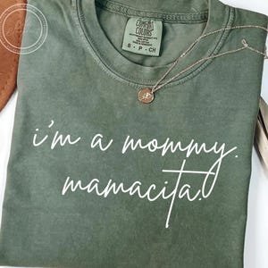 May include: Olive green t-shirt with the text "i'm a mommy. mamacita." in white script. A gold-colored necklace with a circular pendant rests on the shirt. The shirt has a Comfort Colors tag.