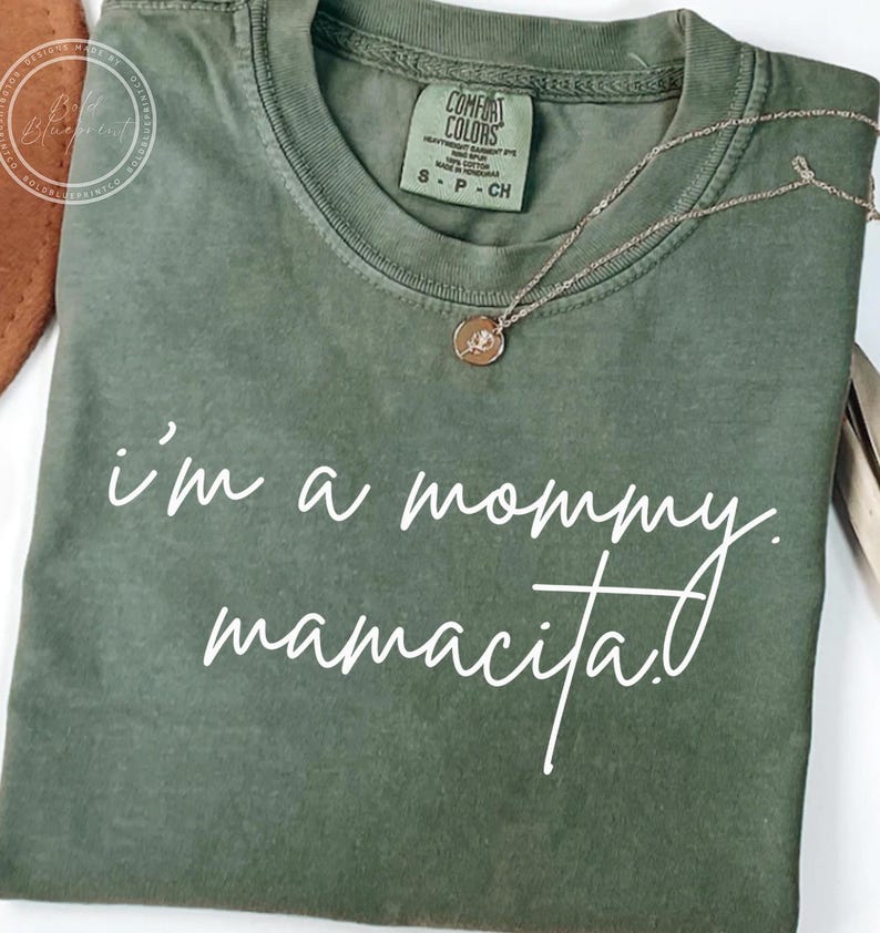 May include: Olive green t-shirt with the text "i'm a mommy. mamacita." in white script. A gold-colored necklace with a circular pendant rests on the shirt. The shirt has a Comfort Colors tag.