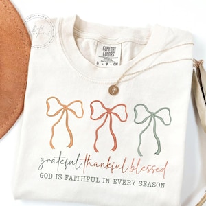 May include: Cream-colored t-shirt with three pastel-colored bow illustrations. The text "grateful thankful blessed" and "GOD IS FAITHFUL IN EVERY SEASON" are printed below the bows. A gold necklace with a pendant is draped on the shirt.
