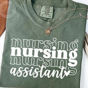 Puede incluir: Una camiseta verde oliva con las palabras "nursing nursing assistant" en blanco, texto apilado. Un collar dorado con un colgante descansa sobre la camisa. La camisa está doblada y tiene una etiqueta Comfort Colors.