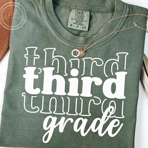 May include: Olive green t-shirt with the words "third grade" in large white letters. A delicate gold necklace with two small pendants rests on the shirt. The shirt has a Comfort Colors tag.