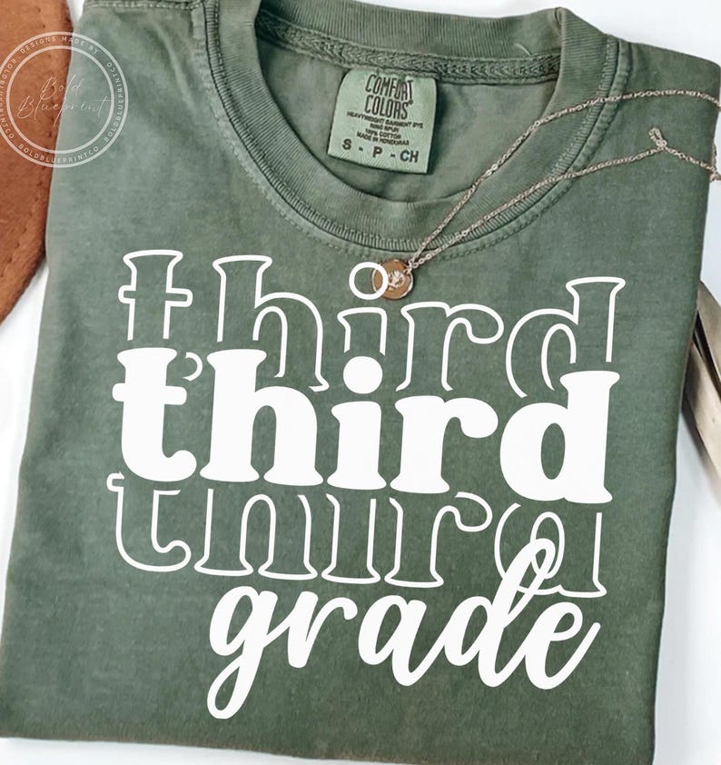 May include: Olive green t-shirt with the words "third grade" in large white letters. A delicate gold necklace with two small pendants rests on the shirt. The shirt has a Comfort Colors tag.
