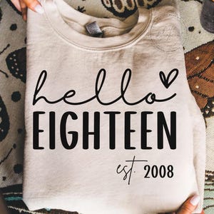 May include: A beige sweatshirt with the words "hello EIGHTEEN" in black script and block letters, with a small heart symbol. Below the text is "est. 2008". The sweatshirt is held by a person.